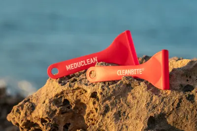 Dos espátulas, una roja con la marca MEDUCLEAN y otra con la marca CLEANBITE, sobre una roca con el mar de fondo.